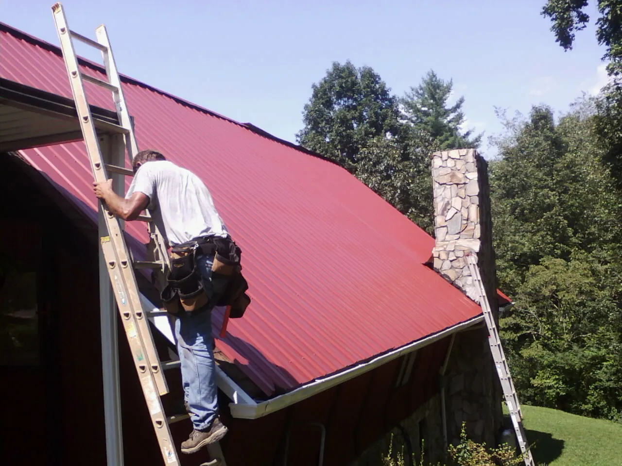 Roofer on a ladder beside a red metal roof and stone chimney — Soffit & Fascia Repair by Roofing Brothers in Grand Ronde
